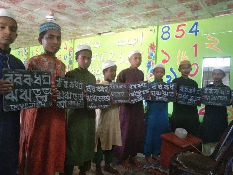 Pupils holding slates showing Bengali handwriting practice in a colourful classroom